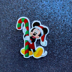 May include: A colorful Christmas-themed sticker featuring Mickey Mouse in a Santa hat, holding a candy cane. Mickey is wearing red shorts, a black shirt, and yellow shoes. The background is a dark, glittery surface.