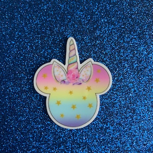 Peut inclure: Une tête de licorne colorée avec une crinière et une corne arc-en-ciel, avec des étoiles dorées sur un fond bleu clair. La tête de licorne est en forme d'oreilles de Mickey Mouse.