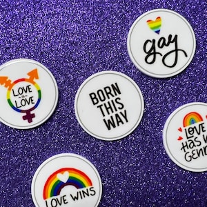 May include: Five white round buttons with rainbow and heart designs and text. The buttons read "Love is Love", "Born This Way", "Love Has No Gender", "Love Wins", and "Gay".