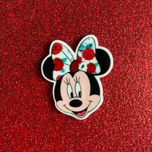 Peut inclure: Un personnage de Minnie Mouse blanc avec un nœud floral rouge et blanc. Le personnage a des oreilles noires, des yeux noirs, un nez noir et une bouche rose. L'arrière-plan est en paillettes rouges.