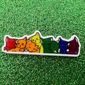 May include: A rainbow-coloured sticker of cartoon cats. The cats are arranged in a row and are red, orange, yellow, green, blue, and purple. The sticker has a white border and is on a green glitter background.