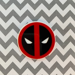 May include: A round, white sticker with a red and black Deadpool logo. The logo features a red outline with a black interior and two white eyes.