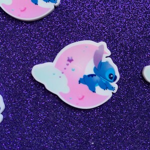 May include: Blue Stitch character sleeping on a pink crescent moon with white clouds and stars.