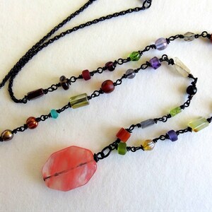May include: A colorful necklace featuring a variety of beads in shades of red, green, purple, and clear, strung on a black chain. The focal point is a large, pink, oval-shaped bead. A unique piece of jewelry.