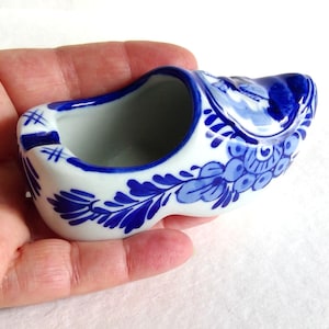 May include: A small, white ceramic clog with blue floral designs. The miniature shoe features a hollow interior and is decorated with intricate blue patterns. The clog is held in a hand, showcasing its size and detail.