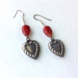 May include: A pair of silver earrings with a heart-shaped pendant and a red teardrop-shaped bead. The heart pendant has a textured border. The earrings hang from silver hooks.