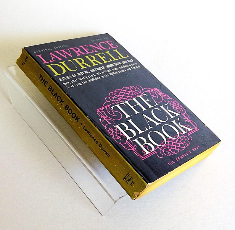 May include: A vintage paperback book titled "The Black Book" by Lawrence Durrell. The cover is black with pink and white text, and a gold spine. The book is in a clear protective sleeve.