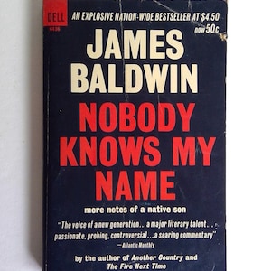 May include: A vintage paperback book titled "Nobody Knows My Name" by James Baldwin. The cover is dark blue with bold white and red lettering. The book is a Dell edition, with text that reads "An Explosive Nation-Wide Bestseller".