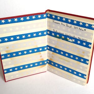 Puede incluir: Libro vintage abierto con cubierta roja y p&aacute;ginas con dise&ntilde;o de rayas azules y blancas con estrellas doradas y blancas. El texto manuscrito en la p&aacute;gina derecha dice "Frances Ann Boyd 309 Palm" y "Bellingham Washington PH 4995".