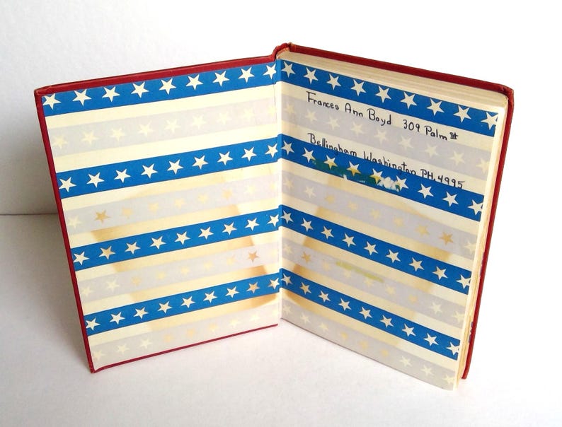 Puede incluir: Libro vintage abierto con cubierta roja y p&aacute;ginas con dise&ntilde;o de rayas azules y blancas con estrellas doradas y blancas. El texto manuscrito en la p&aacute;gina derecha dice "Frances Ann Boyd 309 Palm" y "Bellingham Washington PH 4995".