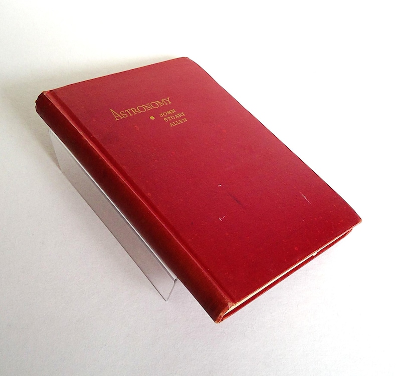 Puede incluir: Un libro de tapa dura rojo titulado "Astronom&iacute;a" con letras doradas. El nombre del autor, John Stuart Allen, tambi&eacute;n est&aacute; impreso en oro. El libro descansa sobre un soporte transparente y en &aacute;ngulo.
