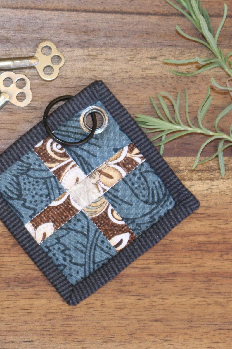 May include: A small, blue and brown patchwork fabric keyring with a black metal ring and a silver metal loop. The keyring is on a wooden surface.