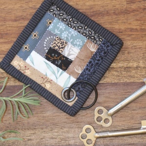 Peut inclure: Un dessous de verre carr&eacute; fait &agrave; la main avec un motif patchwork dans les tons de bleu, marron et cr&egrave;me. Le dessous de verre a une bordure gris fonc&eacute; et un anneau m&eacute;tallique. Deux cl&eacute;s squelettes vintage sont &eacute;galement visibles.