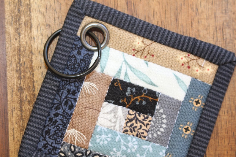Peut inclure: Un petit dessous de verre carr&eacute; matelass&eacute; avec un motif patchwork dans les tons de bleu, marron et cr&egrave;me. Le dessous de verre a une bordure gris fonc&eacute; et un anneau m&eacute;tallique pour suspendre. Le patchwork pr&eacute;sente des motifs floraux et g&eacute;om&eacute;triques.