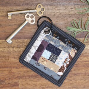 May include: A small, square, quilted keychain with a patchwork design in shades of blue, brown, and beige. The keychain has a black metal ring attached to it.