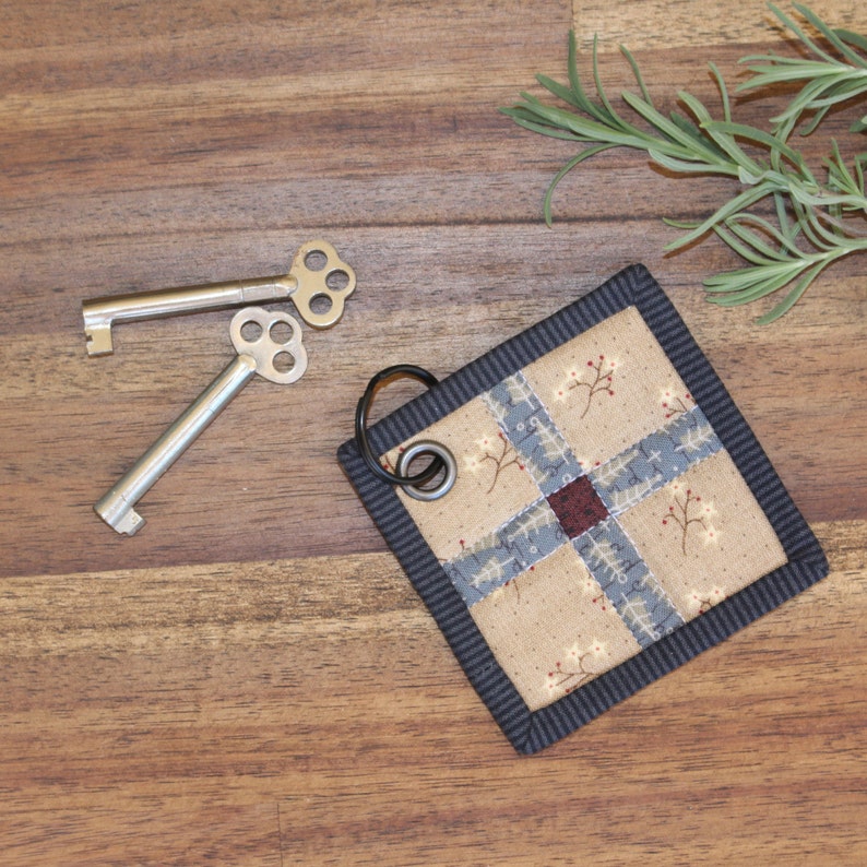 K&ouml;nnte beinhalten: Ein kleiner, quadratischer Schl&uuml;sselanh&auml;nger mit einem blauen und beigefarbenen Patchwork-Design. Der Anh&auml;nger hat einen schwarzen Rand und einen silbernen Metallring. Zwei silberne Schl&uuml;ssel sind ebenfalls auf dem Bild zu sehen.