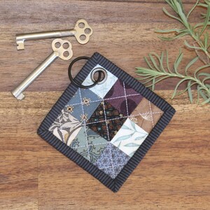 May include: A small, square key fob made of patchwork fabric in shades of blue, brown, and white. The fabric is quilted and has a black border. A silver key ring is attached to the fob.