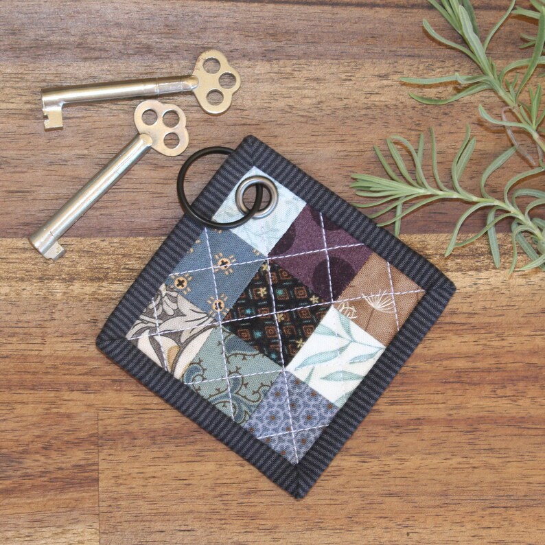May include: A small, square key fob made of patchwork fabric in shades of blue, brown, and white. The fabric is quilted and has a black border. A silver key ring is attached to the fob.