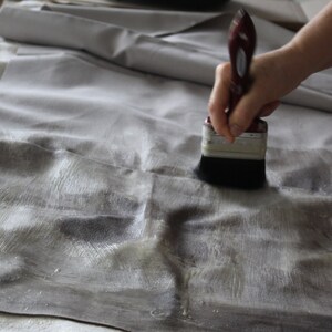 May include: A person's hand holding a paintbrush and applying a white substance to a gray fabric surface.