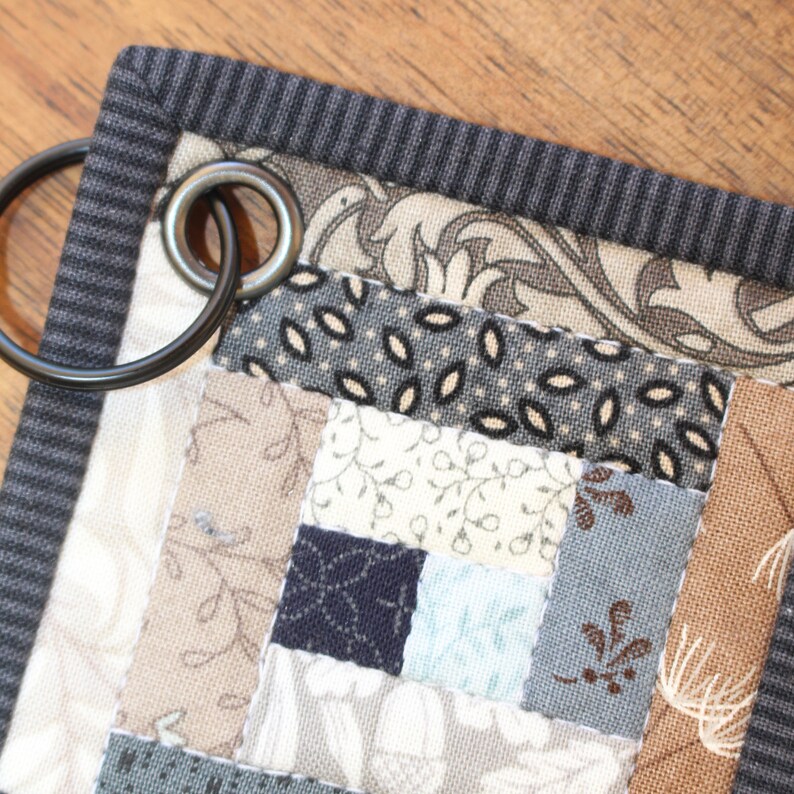 Może przedstawiać: Zbliżenie na szarą i brązową patchworkową kołdrę z czarno-białym wzorem kwiatowym. Kołdra ma czarną obw&oacute;dkę z tkaniny i metalowy pierścień przymocowany do g&oacute;rnego rogu.