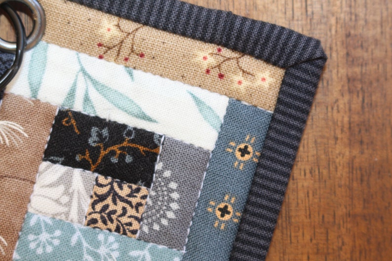 Peut inclure: Gros plan sur un dessous de verre patchwork fait &agrave; la main avec divers motifs de tissu. Le dessous de verre pr&eacute;sente une bordure ray&eacute;e gris fonc&eacute; et un m&eacute;lange de motifs floraux et g&eacute;om&eacute;triques dans des tons de marron, de bleu et de gris. Un anneau m&eacute;tallique est visible.