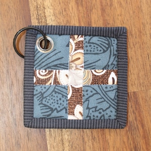 May include: A small, square, grey and brown fabric key chain with a metal ring and a grommet. The fabric is patterned with a cross-shaped design.