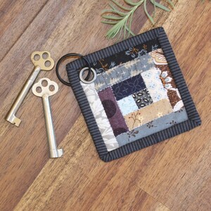 May include: A small, square, patchwork quilt keychain with a black border and a silver key ring. The quilt is made of various fabrics in shades of blue, brown, and purple.