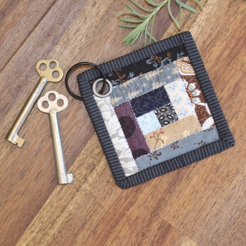 May include: A small, square, patchwork quilt keychain with a black border and a silver key ring. The quilt is made of various fabrics in shades of blue, brown, and purple.