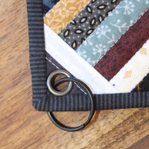 May include: A black metal key ring with a silver ring attached to a gray and white patchwork quilt.