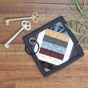 May include: A small, quilted key chain with a black and white striped border. The center of the key chain is made of colorful fabric scraps sewn together in a geometric pattern. A black key ring is attached to the key chain.