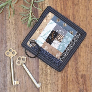 Peut inclure: Un porte-cl&eacute;s matelass&eacute; fait &agrave; la main avec un motif patchwork aux couleurs vari&eacute;es, notamment beige, bleu et gris, attach&eacute; &agrave; un anneau en m&eacute;tal noir. Deux cl&eacute;s squelettes antiques reposent sur une surface en bois, &agrave; c&ocirc;t&eacute; d'une brindille de verdure.