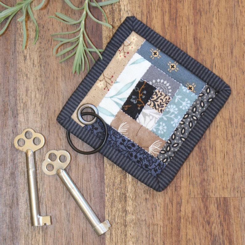 Peut inclure: Un porte-cl&eacute;s matelass&eacute; fait &agrave; la main avec un motif patchwork aux couleurs vari&eacute;es, notamment beige, bleu et gris, attach&eacute; &agrave; un anneau en m&eacute;tal noir. Deux cl&eacute;s squelettes antiques reposent sur une surface en bois, &agrave; c&ocirc;t&eacute; d'une brindille de verdure.