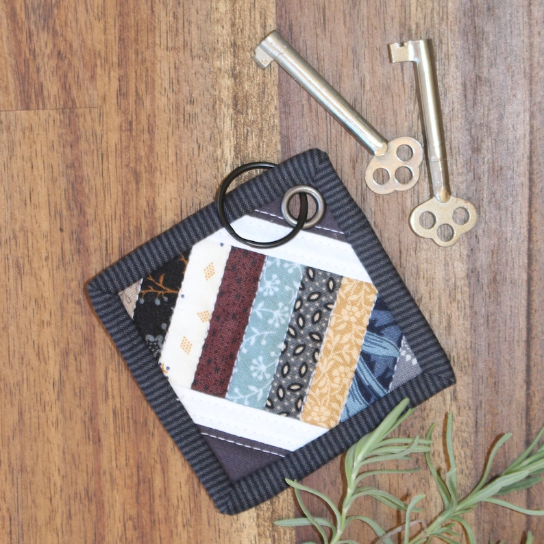 May include: A small, square, quilted key fob with a black border and a patchwork design of various fabrics in shades of brown, blue, and yellow. The fob has a black loop for attaching keys.