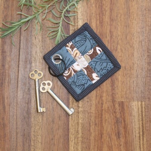 May include: A blue and brown patchwork fabric key holder with a black border and a metal ring. Two antique-style keys are lying on a wooden surface.