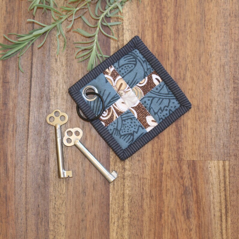 May include: A blue and brown patchwork fabric key holder with a black border and a metal ring. Two antique-style keys are lying on a wooden surface.