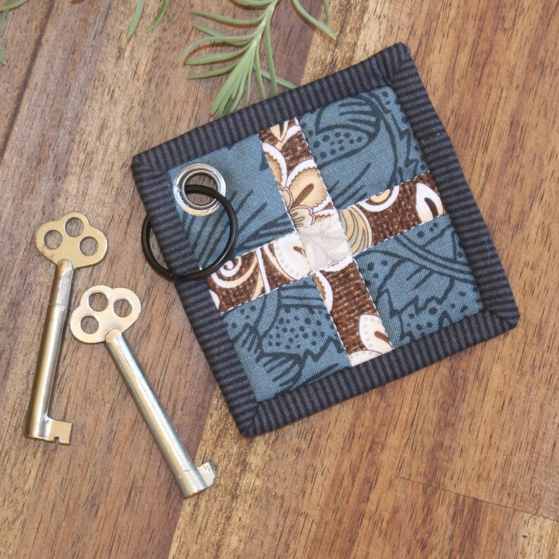 May include: A small, square key fob with a patchwork design of blue, brown and white fabric. The fob has a black border and a metal ring for attaching keys.