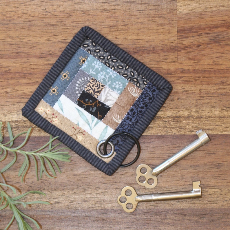 Peut inclure: Un dessous de verre carr&eacute; fait &agrave; la main avec un motif patchwork dans les tons de bleu, beige et marron, avec une bordure gris fonc&eacute;. Deux cl&eacute;s squelettes de style antique et un anneau m&eacute;tallique sont &agrave; c&ocirc;t&eacute; du dessous de verre.