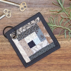 Może przedstawiać: Mały, kwadratowy brelok do kluczy w stylu patchwork z przymocowanym k&oacute;łkiem na klucze. Kołdra wykonana jest z tkanin w r&oacute;żnych odcieniach brązu, szarości i błękitu, z czarno-białą wzorzystą obw&oacute;dką.