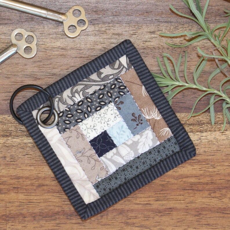 Może przedstawiać: Mały, kwadratowy brelok do kluczy w stylu patchwork z przymocowanym k&oacute;łkiem na klucze. Kołdra wykonana jest z tkanin w r&oacute;żnych odcieniach brązu, szarości i błękitu, z czarno-białą wzorzystą obw&oacute;dką.