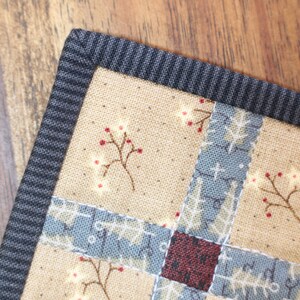 K&ouml;nnte beinhalten: Nahaufnahme einer Stoff-Patchwork-Decke mit schwarzem Rand. Die Decke zeigt verschiedene Muster, darunter ein Blumendesign mit roten Beeren und ein geometrisches Muster in Blau und Wei&szlig;.