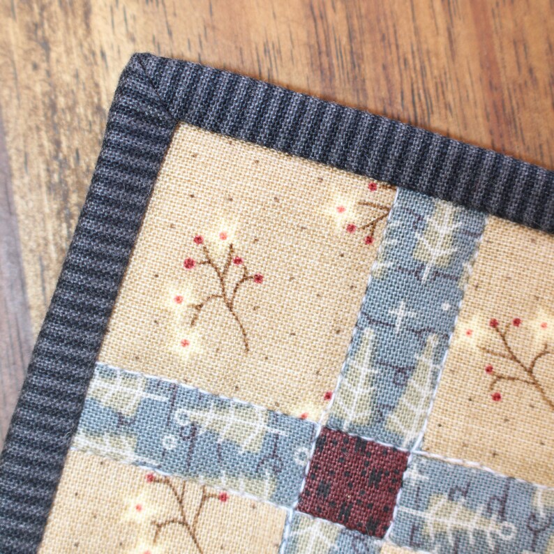 K&ouml;nnte beinhalten: Nahaufnahme einer Stoff-Patchwork-Decke mit schwarzem Rand. Die Decke zeigt verschiedene Muster, darunter ein Blumendesign mit roten Beeren und ein geometrisches Muster in Blau und Wei&szlig;.