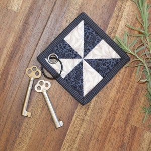 May include: A black and white fabric square with a pinwheel design. The fabric is quilted and has a black border. Two silver keys are on a wooden surface next to the fabric square.