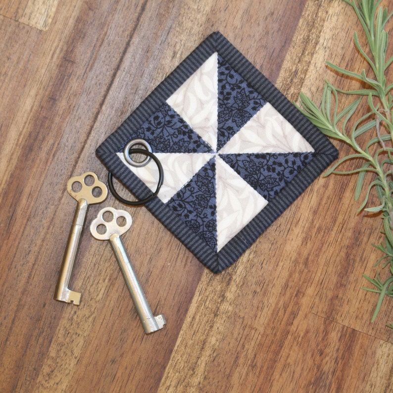 May include: A black and white fabric square with a pinwheel design. The fabric is quilted and has a black border. Two silver keys are on a wooden surface next to the fabric square.