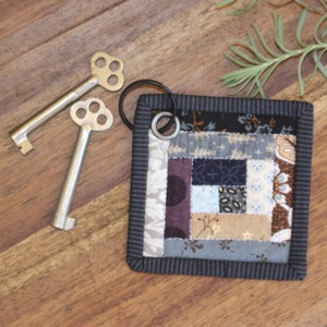 May include: A small, square, patchwork quilt keychain with a black and white striped border. The keychain has a silver key ring attached to it. The keychain is made of various fabrics in shades of brown, gray, purple, and white.