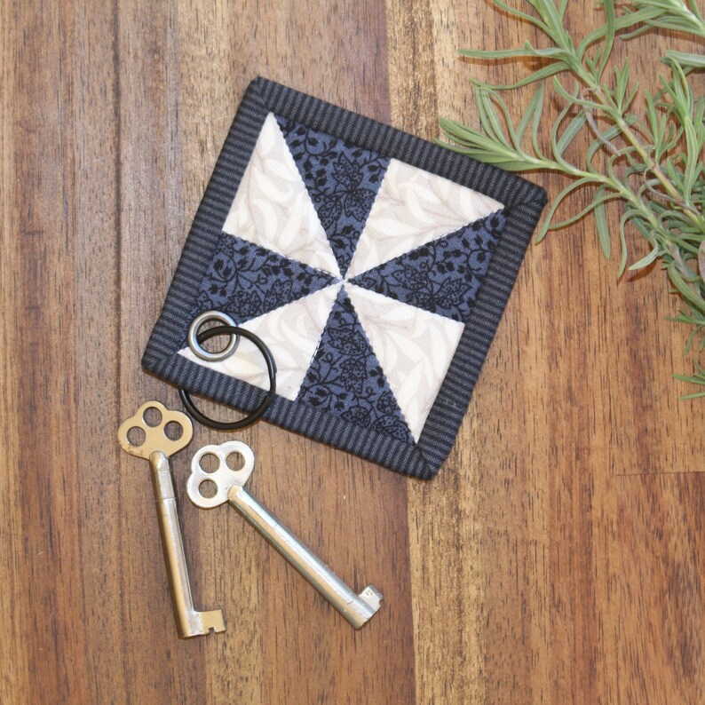May include: A black and white fabric coaster with a pinwheel design. The coaster has a black border and is made of quilted fabric. Two antique keys are lying on a wooden surface next to the coaster.
