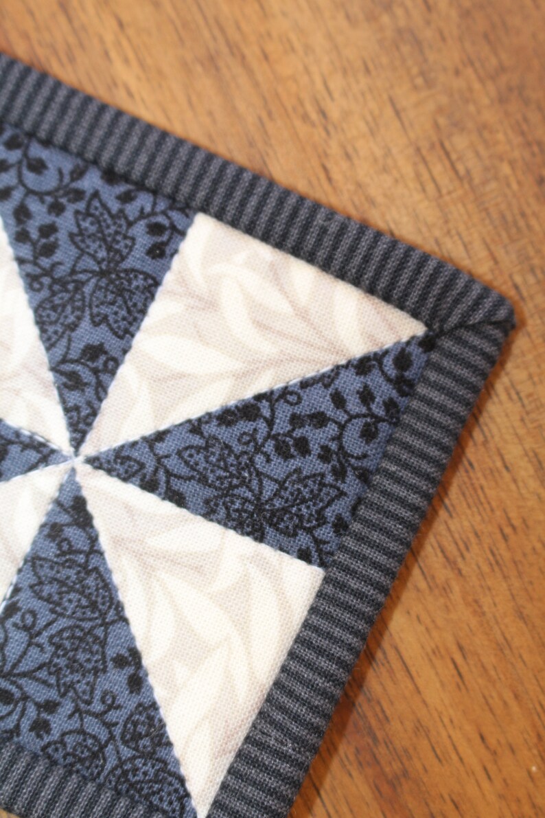 May include: A close-up of a blue and white patchwork quilt square with a black and white floral pattern. The square is surrounded by a grey ribbed fabric border.