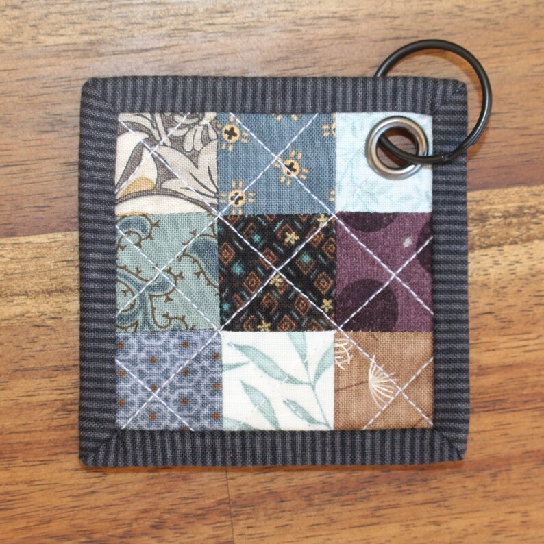 May include: A small, grey, quilted key chain with a variety of fabric patterns in shades of blue, brown, and white. The key chain has a metal ring and a metal grommet.