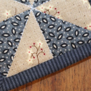 Puede incluir: Primer plano de un posavasos acolchado hecho a mano con un patr&oacute;n de tela azul y blanco y una tela marr&oacute;n con un peque&ntilde;o dise&ntilde;o de &aacute;rbol rojo y blanco. El posavasos tiene un borde de tela acanalada azul oscuro.