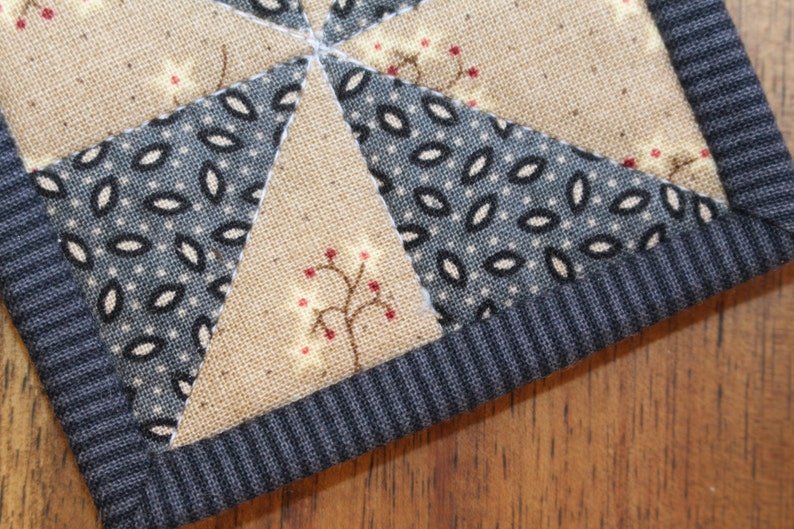 Puede incluir: Primer plano de un posavasos acolchado hecho a mano con un patr&oacute;n de tela azul y blanco y una tela marr&oacute;n con un peque&ntilde;o dise&ntilde;o de &aacute;rbol rojo y blanco. El posavasos tiene un borde de tela acanalada azul oscuro.