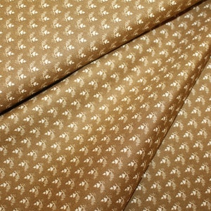 Peut inclure: Gros plan sur un tissu plié de couleur marron chaud, orné d'un motif répétitif. Le motif est composé de petites formes blanches en forme de feuilles stylisées, disposées en grille. Le tissu a une texture subtile.